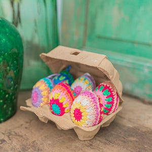 Op de afbeelding: Zes kleurrijke gehaakte paaseieren in een kartonnen eierdoos. De eieren zijn versierd met roze, gele, blauwe en groene garen.