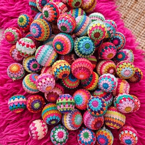 Op de afbeelding: Een verzameling kleurrijke gehaakte ornamenten in verschillende patronen en kleuren, waaronder rood, groen, blauw, geel en roze. De ornamenten zijn op een roze pluche oppervlak in een stapel gelegd.