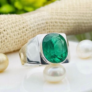 Puede incluir: Un anillo de plata con una gran esmeralda de talla ovalada. La esmeralda es de color verde vibrante y está engastada en una banda rectangular de plata. El anillo se exhibe con perlas.