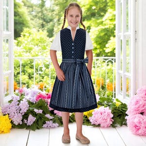 May include: A young girl wearing a blue and white dirndl dress with a white blouse. She is standing in front of a window with flowers on either side.