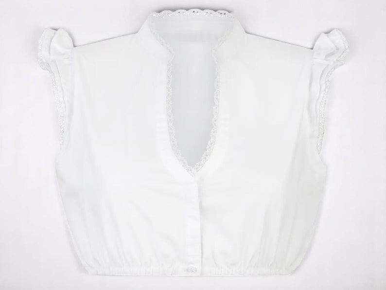 Pu&ograve; includere: Una camicetta bianca senza maniche con colletto alto e arricciato e scollatura a V. La camicetta ha piccoli fiocchi decorativi sulle spalle e una fila di bottoni sul davanti. Il colletto e i bordi delle spalle sono rifiniti con delicato pizzo.