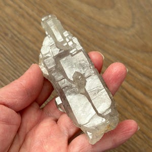 Puede incluir: Un grupo de cristales de cuarzo transparente con una textura natural rugosa. El cristal tiene un aspecto claro y translúcido con estructuras internas visibles. El cuarzo se sostiene en una mano, mostrando su tamaño y forma. El cristal mide aproximadamente 10 cm de largo.