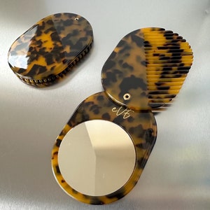 May include: A tortoiseshell compact mirror with a comb on one side and a mirror on the other. The comb has a tortoiseshell design and the mirror is round. The compact is closed and the comb is visible.