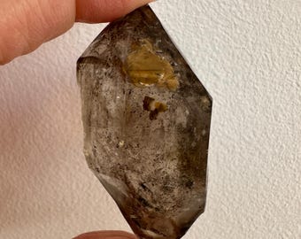 Op de afbeelding: Een gefacetteerde rookkwartskristal, vastgehouden tussen vingers. Het kristal heeft een doorschijnende bruine kleur met donkere insluitsels en een goudbruine vlek. Het kristal heeft een zeshoekige vorm met een punt. De achtergrond is een effen, gebroken witte muur.