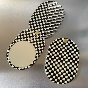 Può includere: Uno specchio compatto a scacchi bianco e nero con un pettine sul retro. Lo specchio è di forma ovale e il pettine è a forma di semicerchio.