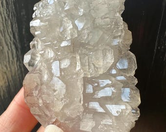Op de afbeelding: Een heldere, kristallijne kwartskluster met meerdere facetten. De natuurlijke minerale formatie heeft een ruw, getextureerd oppervlak en een doorschijnend uiterlijk. De kwarts wordt in een hand gehouden, met een donkere achtergrond.