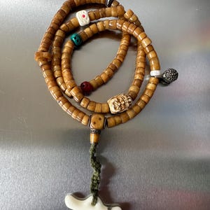 May include: A string of cylindrical tan beads arranged in a circular pattern, accented with a carved white face bead, a small red bead, a turquoise bead, and a dark metal textured charm. A green cord hangs from a square bead to a white bone-like pendant.