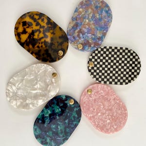 Op de afbeelding: Een verzameling van zes ovale decoratieve trays in verschillende kleuren en patronen. De trays bevatten een schildpadontwerp, een blauw-paars gespikkeld patroon, een zwart-wit dambord, parelmoerwit, een blauwgroen en marineblauw gespikkeld patroon en een roze gespikkeld patroon.