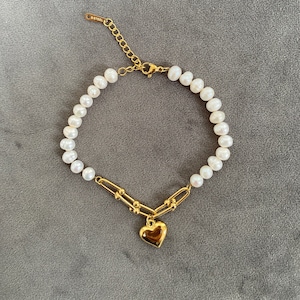 Op de afbeelding: Een gouden kettingarmband met een hartvormige hanger en witte parels. De ketting is gemaakt van kleine, met elkaar verbonden cirkels. De hartvormige hanger is massief en heeft een glad oppervlak. De parels zijn gelijkmatig verdeeld en hebben een uniforme grootte.