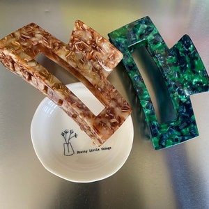 Puede incluir: Dos pinzas grandes para el pelo, una marrón y crema, la otra verde y negra. Están sobre un plato blanco pequeño con el texto "Pretty little things".