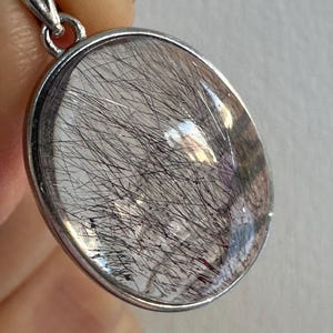 Op de afbeelding: Ovale hanger met een heldere, gepolijste steen met zwarte, haarachtige insluitsels. De steen is gezet in een zilverkleurig metalen frame met een decoratieve beugel. De hanger wordt door een hand omhoog gehouden.