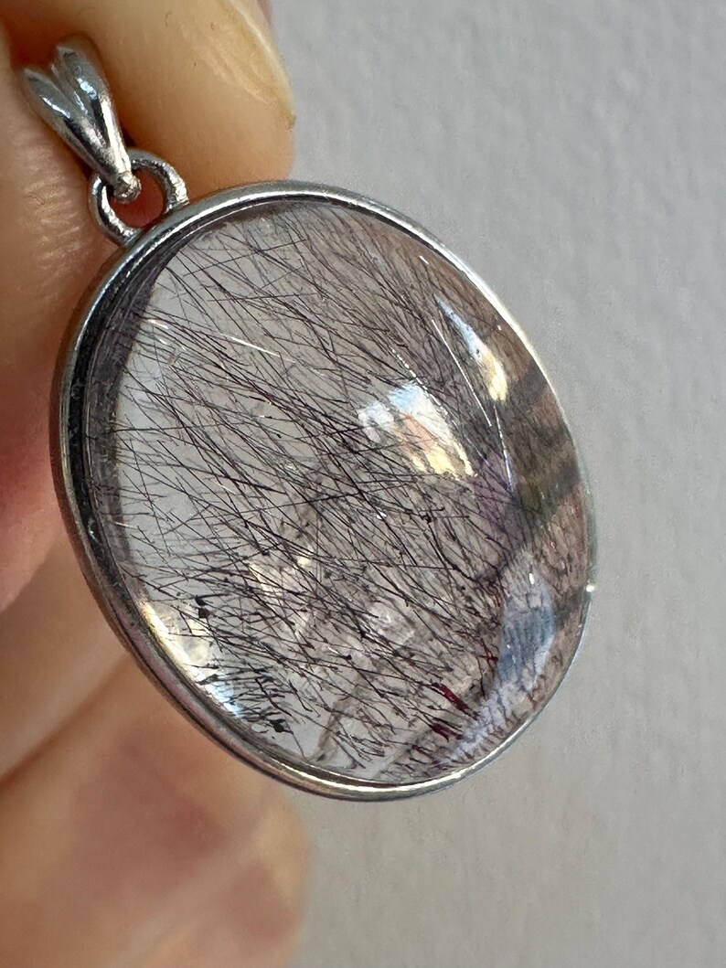 Op de afbeelding: Ovale hanger met een heldere, gepolijste steen met zwarte, haarachtige insluitsels. De steen is gezet in een zilverkleurig metalen frame met een decoratieve beugel. De hanger wordt door een hand omhoog gehouden.