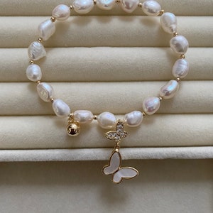 May include: A white pearl bracelet with a gold butterfly charm. The bracelet has a small gold bell charm and is strung on a gold chain.
