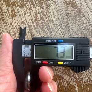 Op de afbeelding: Een digitale schuifmaat meet een klein, rond object. Het digitale display toont een meting van 3 mm. De schuifmaat is zwart met zilveren meetarmen. Het object lijkt een ring of een kleine steen te zijn.