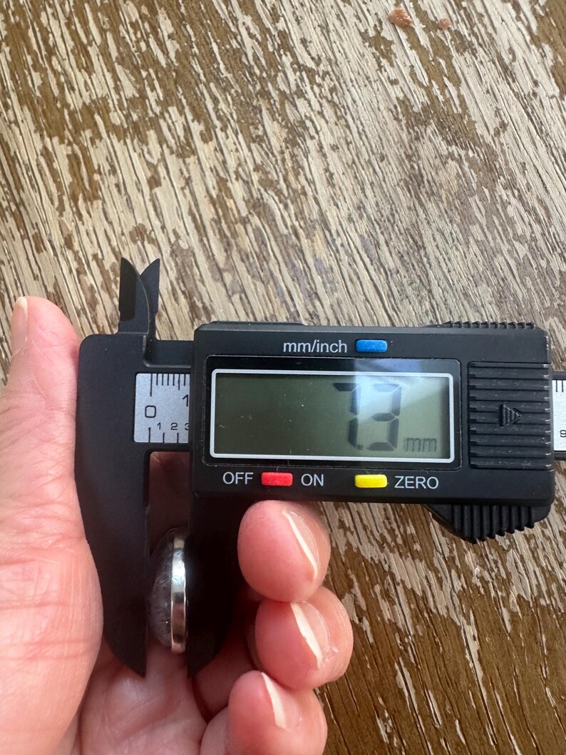 Op de afbeelding: Een digitale schuifmaat meet een klein, rond object. Het digitale display toont een meting van 3 mm. De schuifmaat is zwart met zilveren meetarmen. Het object lijkt een ring of een kleine steen te zijn.