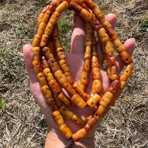 May include: A long strand of amber-colored beads, each carved with a small, stylized face. The beads vary in shades of translucent orange, yellow, and deep brown, strung together on a dark brown cord with a knotted tassel at the bottom end.