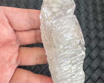 Op de afbeelding: Een heldere, kristallijne kwartskluster met een ruw, getextureerd oppervlak. De kwarts wordt in een hand gehouden, waarbij de natuurlijke, onregelmatige vorm en doorschijnende kwaliteit worden getoond. Het kristal lijkt ongepolijst, wat de ruwe, natuurlijke schoonheid benadrukt.