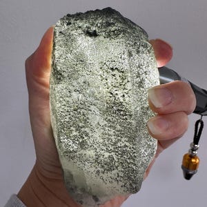 May include: A large, rough mineral specimen with a metallic, golden-flecked surface texture. The stone is translucent, allowing a bright light source to shine through its crystalline structure, highlighting dark, granular inclusions throughout.