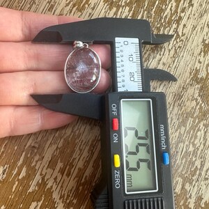 Op de afbeelding: Ovale hanger met een zilverkleurige zetting, met een doorschijnende, roodbruine steen. De hanger wordt gemeten met een digitale schuifmaat, met een afmeting van 2,55 cm.