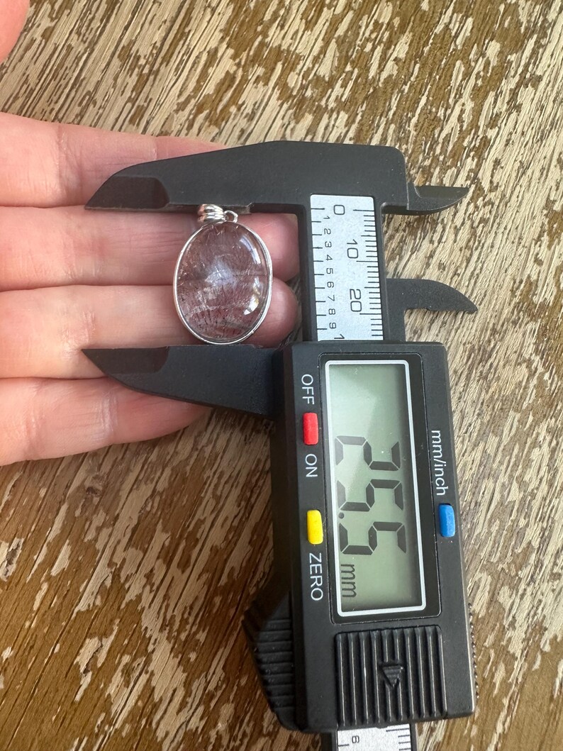 Op de afbeelding: Ovale hanger met een zilverkleurige zetting, met een doorschijnende, roodbruine steen. De hanger wordt gemeten met een digitale schuifmaat, met een afmeting van 2,55 cm.