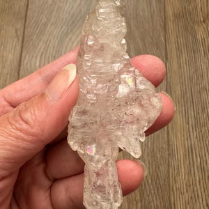 May include: A clear, raw crystal cluster with a rough, textured surface. The crystal has a natural, irregular shape and exhibits subtle iridescent flashes. The crystal is held in a hand against a wooden background.