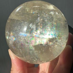 May include: A polished, spherical clear quartz crystal ball featuring internal fractures and inclusions that create vibrant, multi-colored rainbow light refractions throughout the transparent, glass-like surface of the stone.