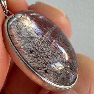 Op de afbeelding: Ovale hanger met een heldere, gepolijste edelsteen met zwarte, haarachtige insluitsels. De steen is gezet in een zilverkleurige metalen rand met een kleine lus voor een ketting. De hanger wordt in een hand gehouden.