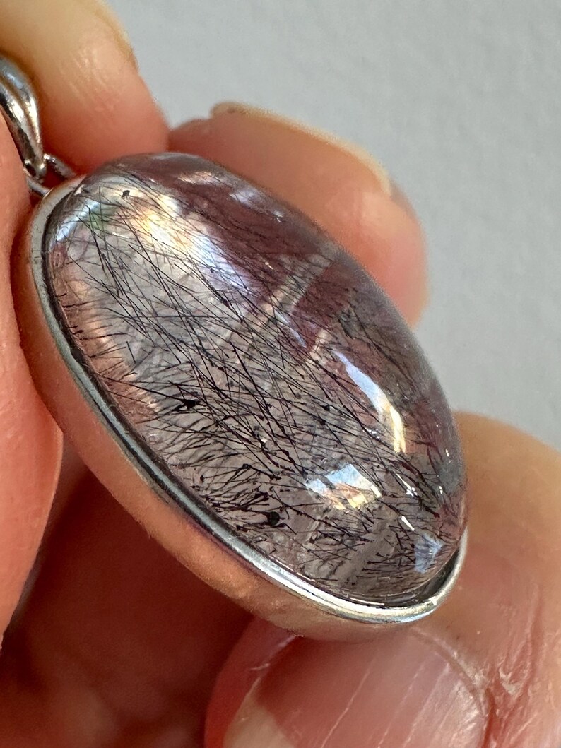 Op de afbeelding: Ovale hanger met een heldere, gepolijste edelsteen met zwarte, haarachtige insluitsels. De steen is gezet in een zilverkleurige metalen rand met een kleine lus voor een ketting. De hanger wordt in een hand gehouden.
