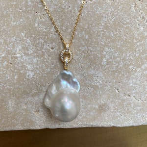 Pu&ograve; includere: Una collana con catena d'oro con un grande ciondolo di perla barocca bianca. Il ciondolo &egrave; attaccato a un piccolo cerchio dorato con pietre chiare e scintillanti.