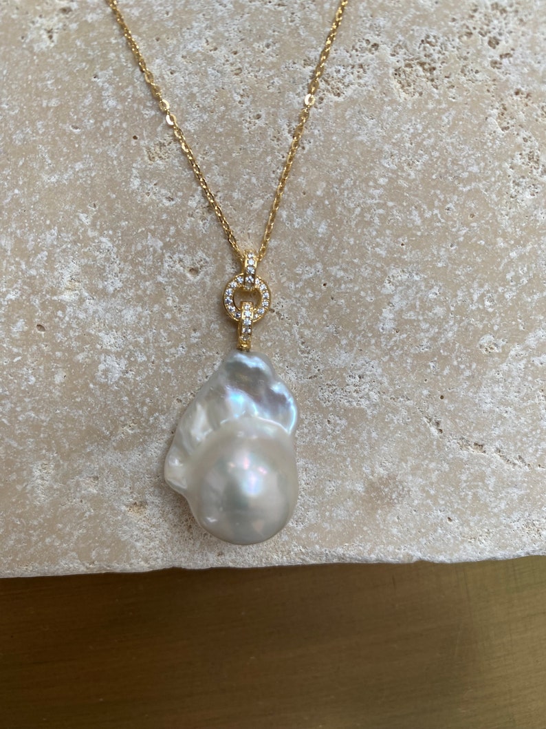 Pu&ograve; includere: Una collana con catena d'oro con un grande ciondolo di perla barocca bianca. Il ciondolo &egrave; attaccato a un piccolo cerchio dorato con pietre chiare e scintillanti.
