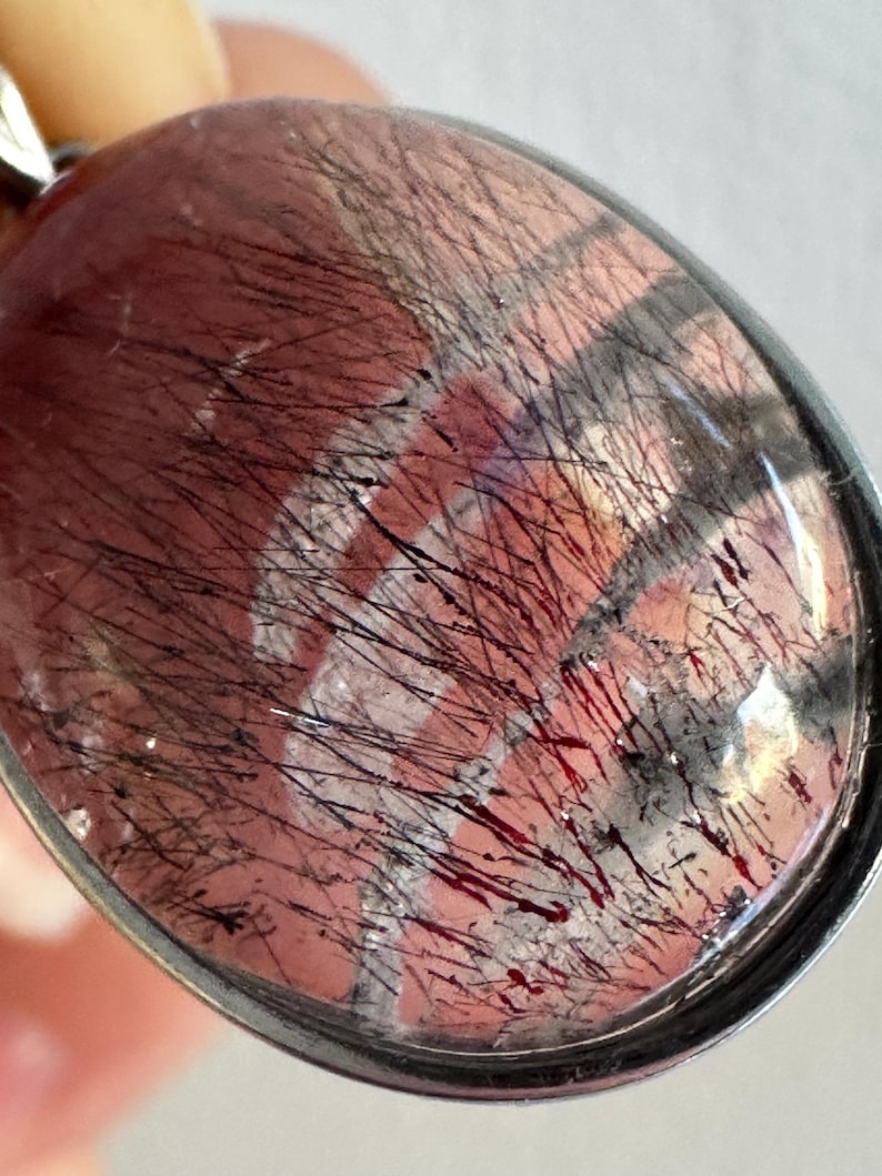 Op de afbeelding: Ovale hanger met een helder, gepolijst oppervlak. De hanger heeft een roodbruine basis met zwarte, haarachtige insluitsels en een zilverkleurig metalen frame. De insluitsels cre&euml;ren een opvallende visuele textuur.