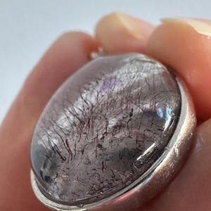 Op de afbeelding: Ovale hanger met een heldere, gepolijste steen met donkerrode en zwarte insluitsels, gezet in een zilverkleurige metalen rand. De steen heeft een uniek, natuurlijk patroon. De hanger wordt in een hand gehouden.