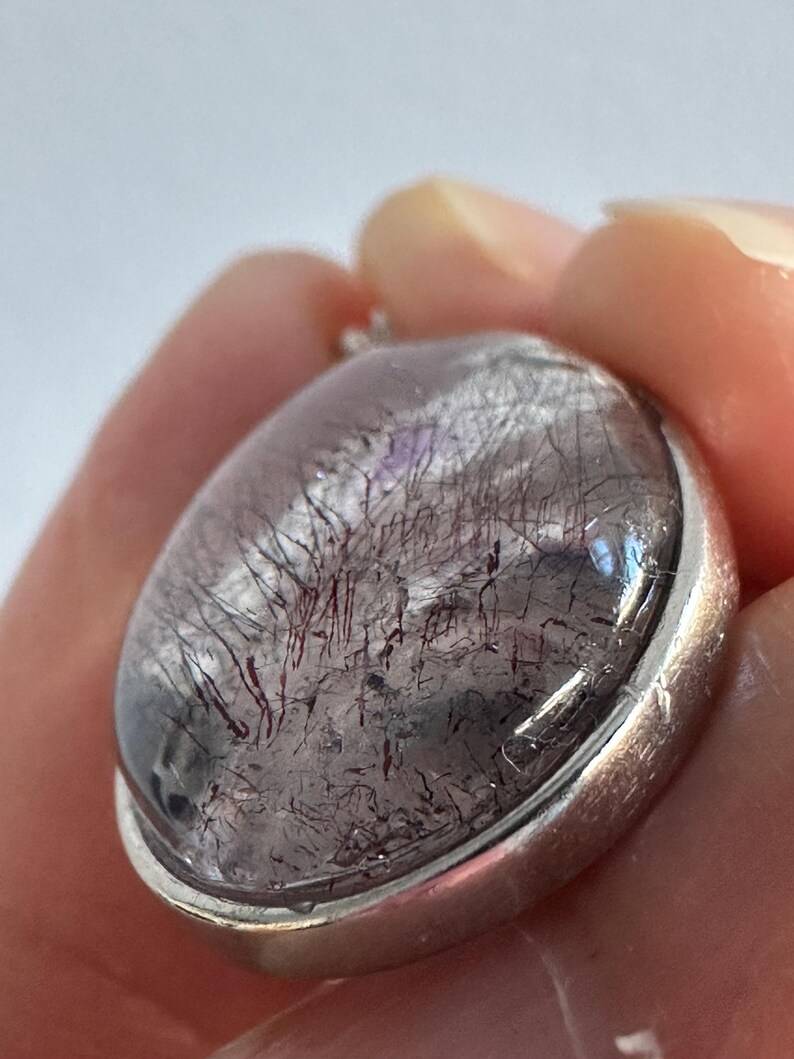 Op de afbeelding: Ovale hanger met een heldere, gepolijste steen met donkerrode en zwarte insluitsels, gezet in een zilverkleurige metalen rand. De steen heeft een uniek, natuurlijk patroon. De hanger wordt in een hand gehouden.