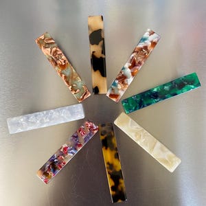 May include: A selection of rectangular hair clips in diverse colours and patterns. The clips feature shades of brown, green, white, and tortoiseshell, with marbled and speckled designs. The clips are arranged in a circular pattern on a silver surface.