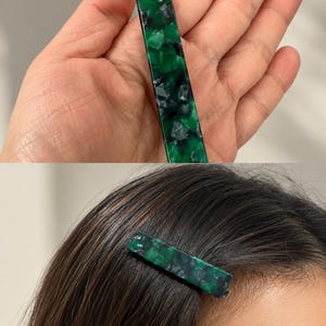 Puede incluir: Una pinza para el pelo rectangular con un patr&oacute;n veteado verde y negro. La pinza se muestra en una mano y tambi&eacute;n en el pelo de una persona. La pinza mide aproximadamente 7,6 cm de largo.