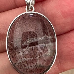 Op de afbeelding: Ovale hanger met een zilverkleurige beugel. De hanger heeft een doorschijnende, roodbruine steen met donkere, haarachtige insluitsels. De steen is gezet in een zilverkleurige bezel.