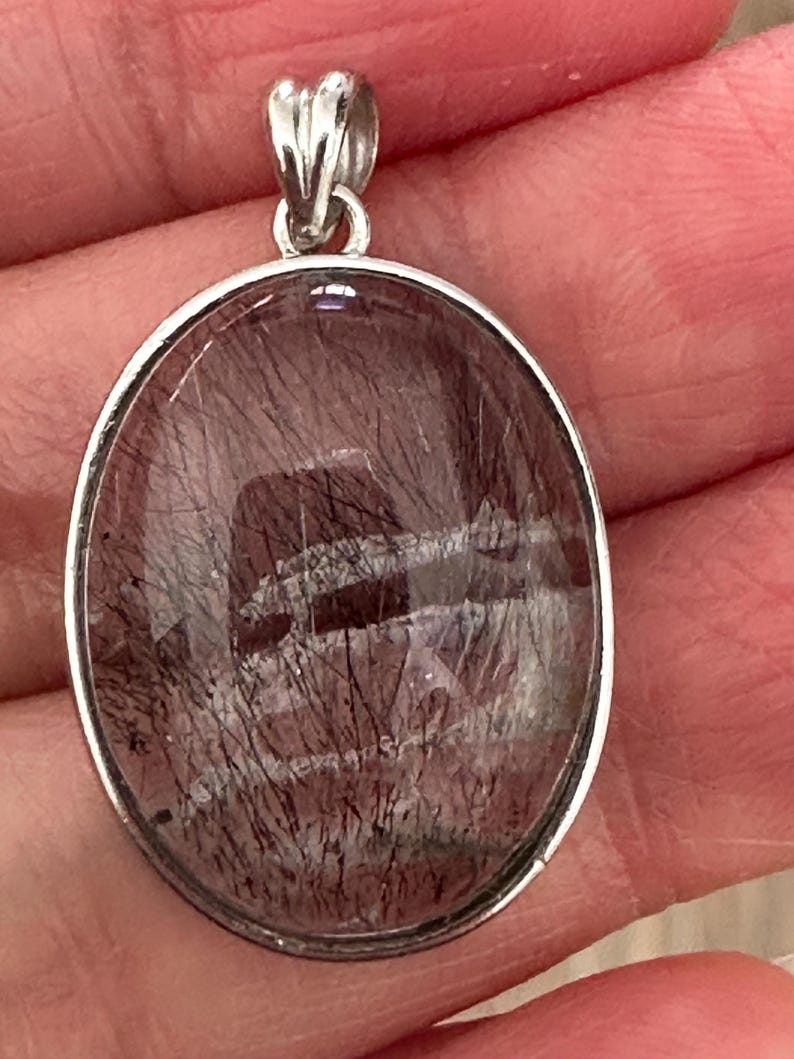 Op de afbeelding: Ovale hanger met een zilverkleurige beugel. De hanger heeft een doorschijnende, roodbruine steen met donkere, haarachtige insluitsels. De steen is gezet in een zilverkleurige bezel.