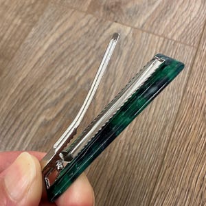 Puede incluir: Una pinza para el pelo de metal plateado con un dise&ntilde;o rectangular veteado en verde oscuro y negro. El clip tiene un mecanismo de bisagra y una serie de dientes para sujetar el cabello. El accesorio para el cabello est&aacute; dise&ntilde;ado para sujetar el cabello en su lugar.