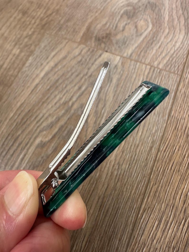 Puede incluir: Una pinza para el pelo de metal plateado con un dise&ntilde;o rectangular veteado en verde oscuro y negro. El clip tiene un mecanismo de bisagra y una serie de dientes para sujetar el cabello. El accesorio para el cabello est&aacute; dise&ntilde;ado para sujetar el cabello en su lugar.