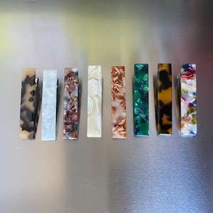 Puede incluir: Una colecci&oacute;n de nueve pinzas para el pelo rectangulares en varios colores y patrones. Las pinzas presentan dise&ntilde;os que incluyen remolinos negros y marrones, m&aacute;rmol blanco y patrones moteados verdes y marrones. Cada pinza mide unos 7.6 cm de largo.