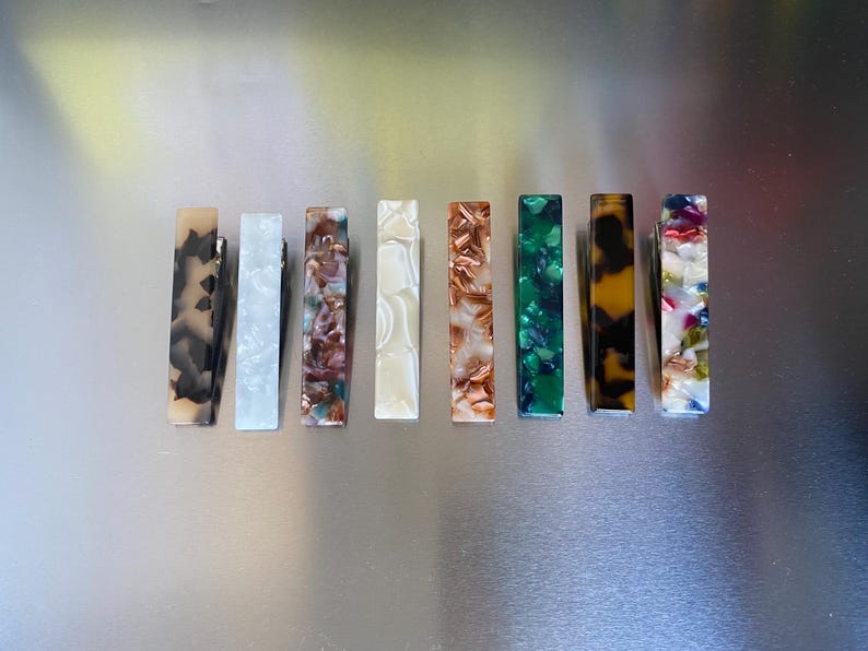 Puede incluir: Una colecci&oacute;n de nueve pinzas para el pelo rectangulares en varios colores y patrones. Las pinzas presentan dise&ntilde;os que incluyen remolinos negros y marrones, m&aacute;rmol blanco y patrones moteados verdes y marrones. Cada pinza mide unos 7.6 cm de largo.