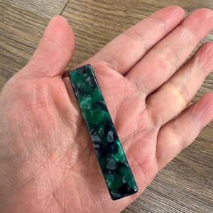 Puede incluir: Una pinza de pelo rectangular con un patr&oacute;n veteado en verde oscuro y negro. La pinza se sostiene en una mano, mostrando su acabado brillante y variaciones de color. El accesorio para el cabello mide aproximadamente 7,6 cm de largo.