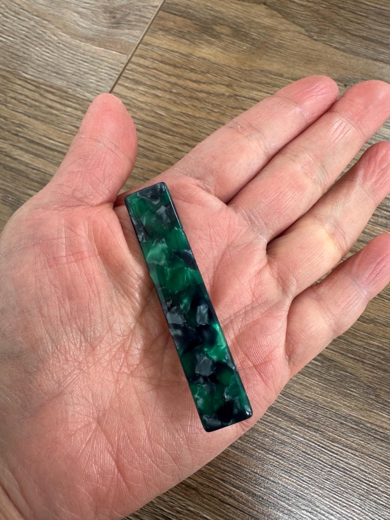 Puede incluir: Una pinza de pelo rectangular con un patr&oacute;n veteado en verde oscuro y negro. La pinza se sostiene en una mano, mostrando su acabado brillante y variaciones de color. El accesorio para el cabello mide aproximadamente 7,6 cm de largo.