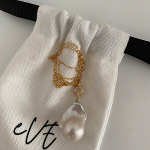 Pu&ograve; includere: Una collana a catena dorata con un grande ciondolo a forma di perla barocca bianca e un piccolo ciondolo con diamanti incastonati. La collana &egrave; in una borsa di stoffa bianca con la scritta "evt jewellery" stampata su di essa.