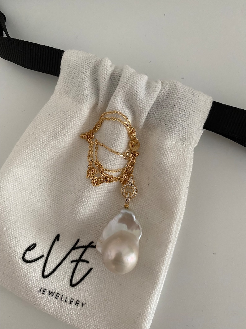 Pu&ograve; includere: Una collana a catena dorata con un grande ciondolo a forma di perla barocca bianca e un piccolo ciondolo con diamanti incastonati. La collana &egrave; in una borsa di stoffa bianca con la scritta "evt jewellery" stampata su di essa.