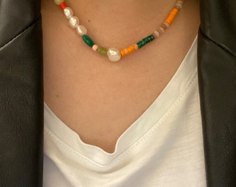 May include: A colorful beaded necklace with a variety of shapes and colors. The necklace features a large pearl in the center, surrounded by green, orange, and pink beads. The necklace is worn over a white t-shirt and black jacket.