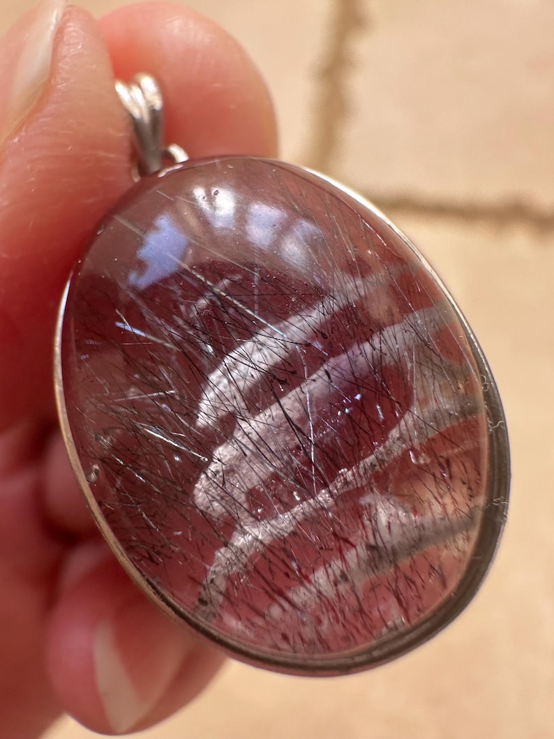 Op de afbeelding: Ovale hanger met een zilverkleurige beugel. De hanger heeft een doorschijnende, roodbruine steen met zwarte en zilveren insluitsels. De steen is gezet in een zilverkleurige bezel.