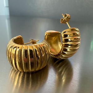 May include: A pair of gold hoop earrings. The earrings feature a ribbed design and a post closure. The earrings are presented on a reflective surface. The earrings are a jewellery accessory.