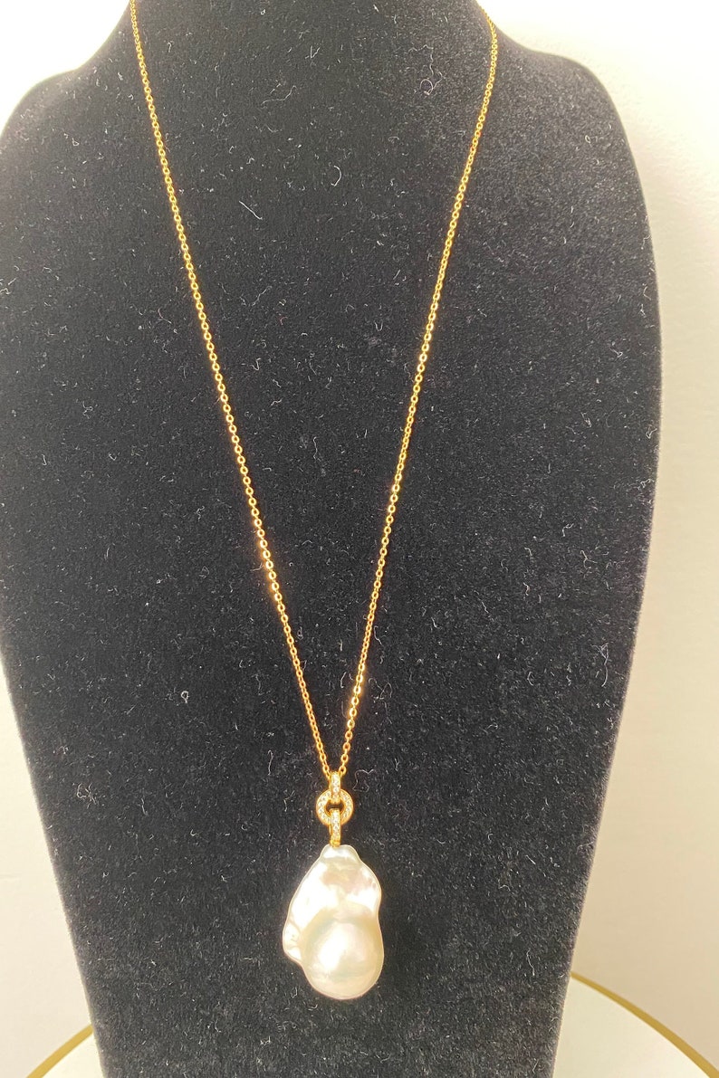 Pu&ograve; includere: Una collana con catena d'oro con un grande ciondolo di perla barocca bianca. La perla &egrave; attaccata alla catena con un piccolo anello d'oro che &egrave; incastonato con piccoli diamanti.