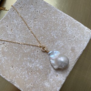 Pu&ograve; includere: Una collana con catena dorata con un grande ciondolo a forma di perla barocca bianca. Il ciondolo &egrave; attaccato alla catena con un piccolo anello dorato che &egrave; incastonato con piccole pietre trasparenti.